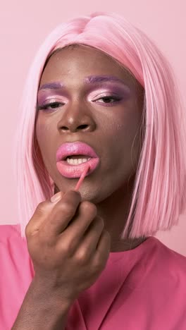 extravagant black transgender applying lip gloss in studio against pink background
