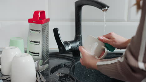 close-up partial view of lady washing dishes with running tap water, scrubbing a cup with sponge, cleaning dishes in modern kitchen sink, household chores, kitchen hygiene