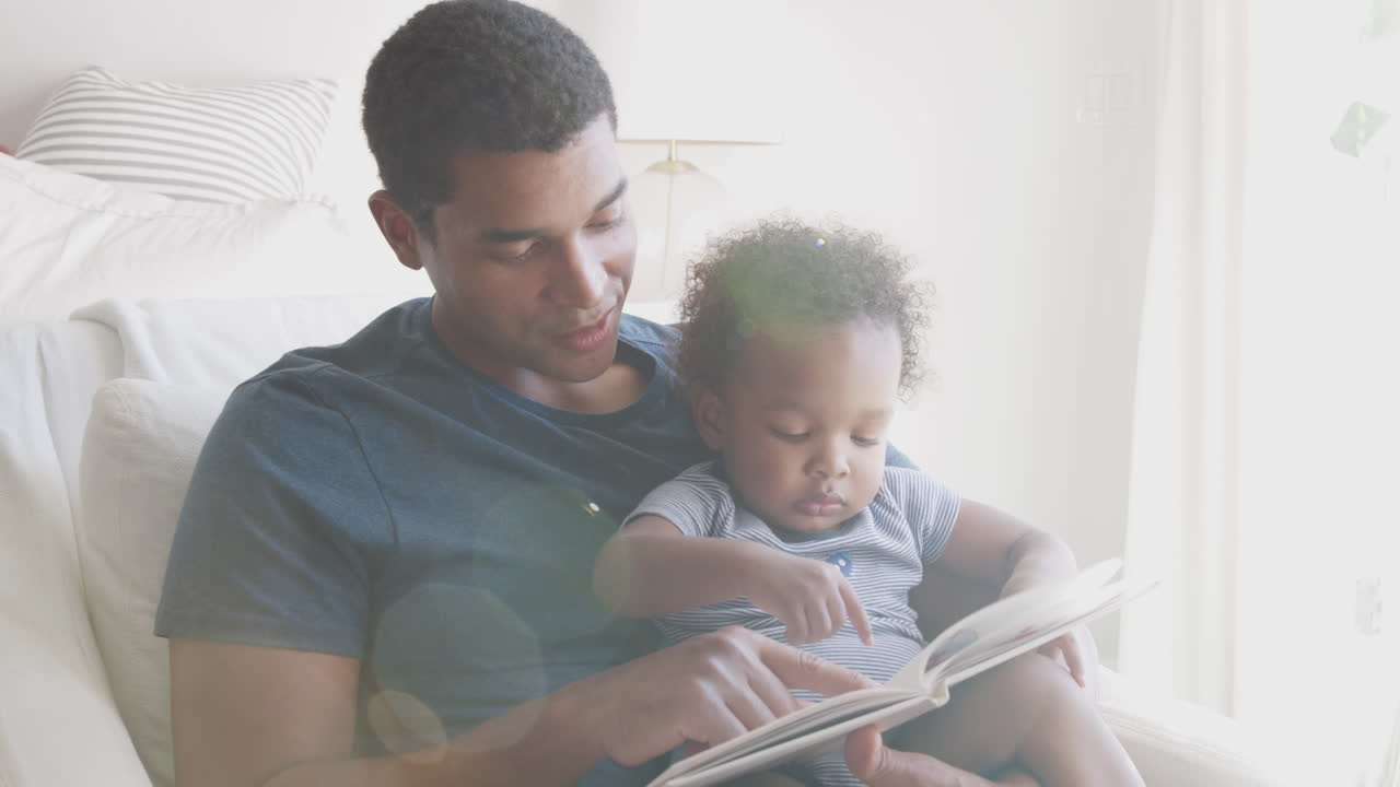 milenial padre afroamericano sentado y leyendo un libro con su hijo pequeño, vista frontal, de cerca