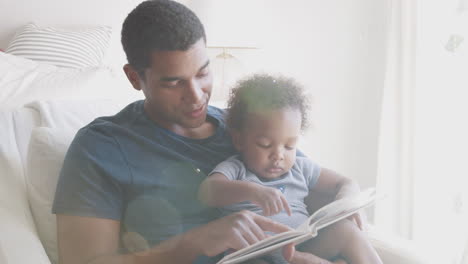 millennial african american father sitting and reading a book with his toddler son, front view, close up