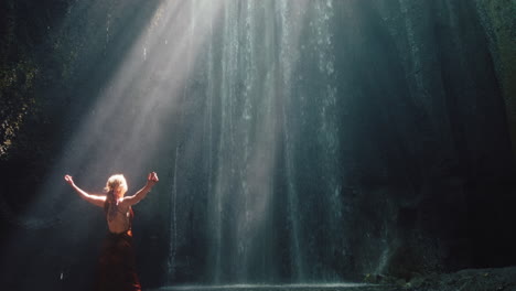 Tanzende-Frau-In-Der-Wasserfallhöhle,-Die-Wasser-Spritzt-Und-Ein-Wunderschönes-Kleid-Trägt,-Genießt-Die-Natur,-Tanzt-Und-Fühlt-Spirituelle-Freiheit-4k