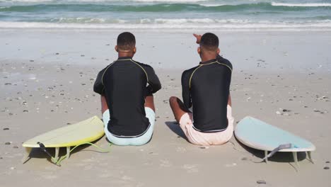 african american teenage twin brothers sitting by surfboards on a beach talking