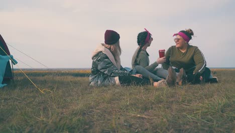 happy women with drinks look at sky sitting on grass by tent