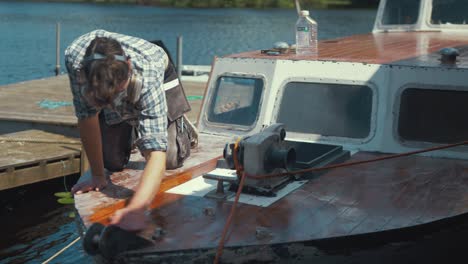 Young-carpenter-wiping-down-dust-off-sanded-wooden-boat-bow-before-varnishing