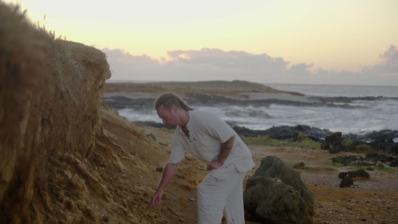 A man in white clothing stands still on a rocky coastal path, glancing around as waves break against the shoreline under warm morning light.