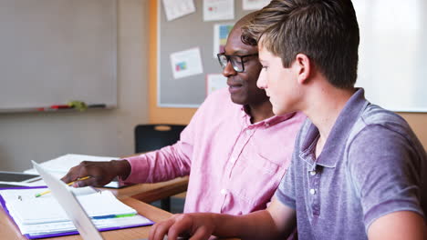 High-School-Tutor-Giving-Male-Student-Using-Laptop-One-To-One-Tuition-At-Desk