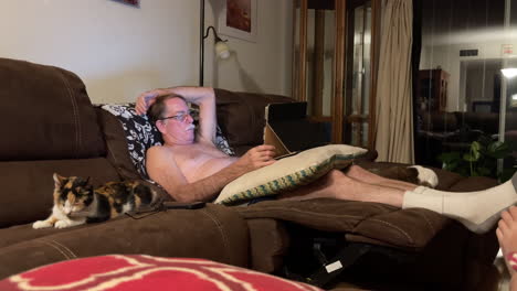 shirtless senior male relaxes on couch with notebook computer, cat next to him