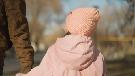 little girl in pink cap and jacket, holding an adult's hand while walking in a sunlit park, with blurred trees and buildings in the background