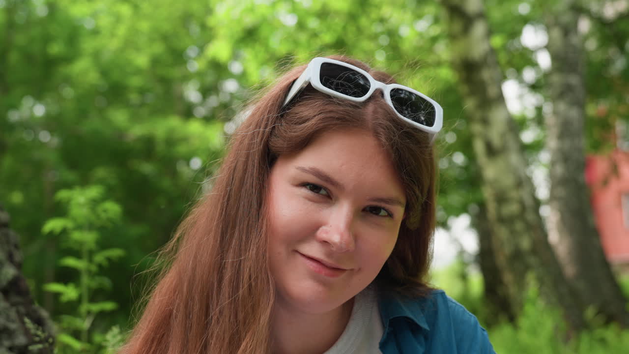 Lovely lady with warm smile wearing white sunglasses on head outdoors in lush green park, surrounded by soft blurred trees and natural light, expressing cheerful mood and gentle confidence