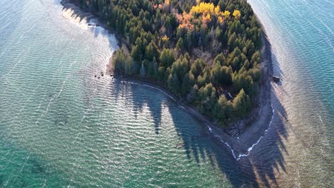 drone footage of a forested peninsula with autumn colors, turquoise shallows, and long tree shadows on a remote shoreline