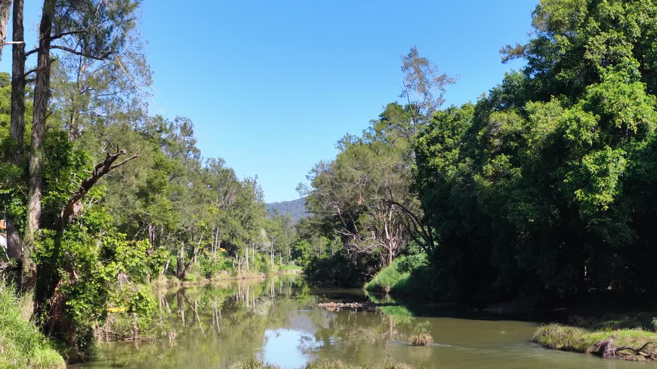 A serene stream flows through lush forests under clear blue skies in Uki, NSW, Australia