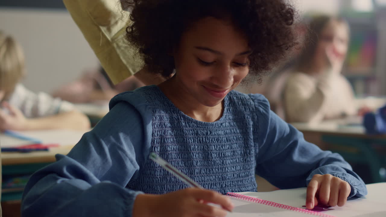 Girl talking with teacher in classroom. Smiling schoolgirl writing in notebook