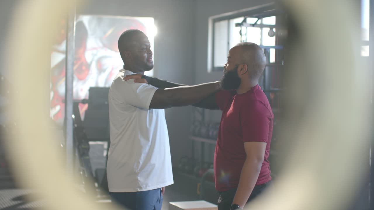 African men trainer in white polo approaching red tee client, assessing posture with weights