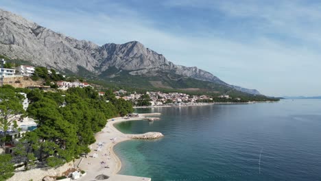 low angle drone brela croatia summers day wispy cloud shady pine trees on beach edge