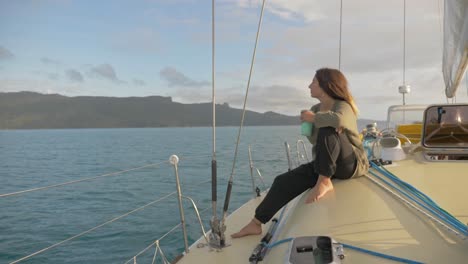 mujer caucásica sentada al costado de un velero, mirando lejos mientras sostiene una taza