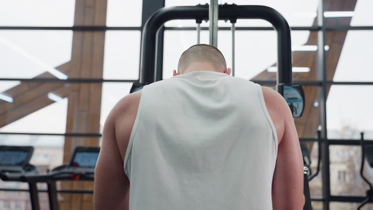 Back view of student with head down pulling chest press machine handle demonstrating focused upper body strength and chest muscles working under modern gym lighting with large windows