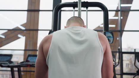 back view of student with head down pulling chest press machine handle demonstrating focused upper body strength and chest muscles working under modern gym lighting with large windows