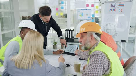 good-looking confident purposeful diverse team of specialists of building industry holding a meeting and discussing details of joint building project when looking at tablet pc with depicted drawing