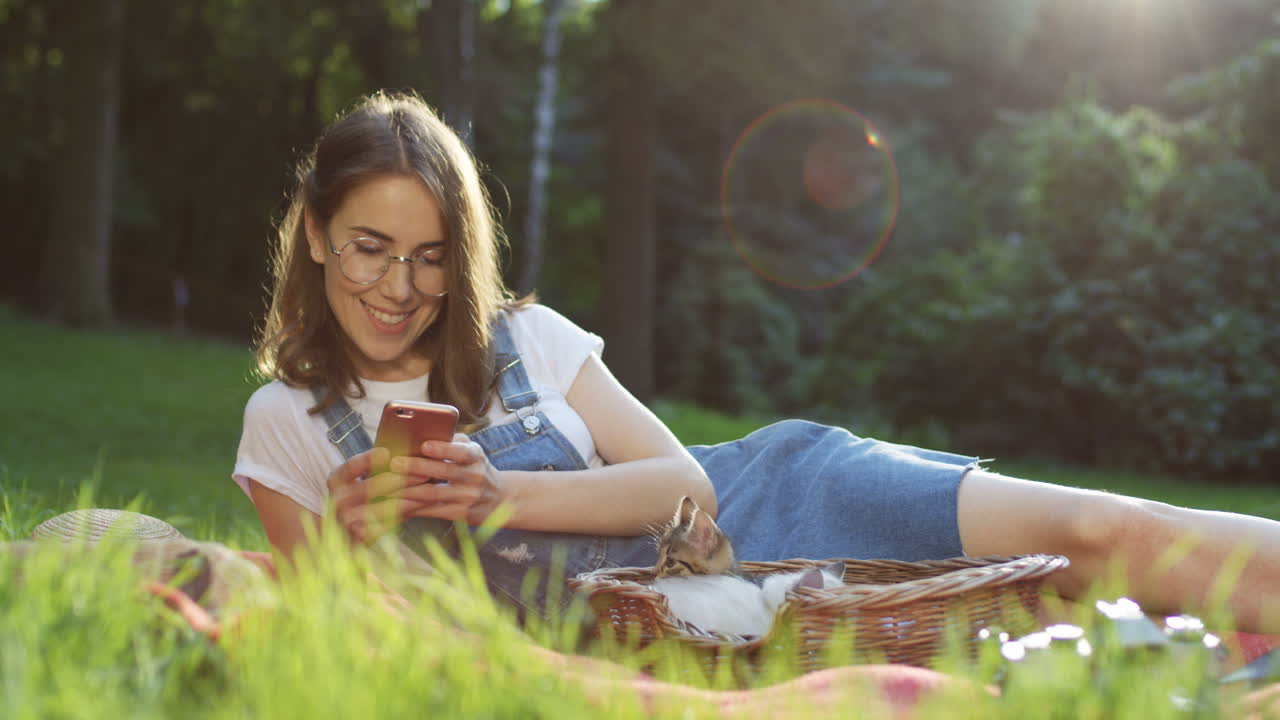 joven mujer caucásica con anteojos acostada sobre una manta en el parque tocando y desplazándose en un teléfono inteligente