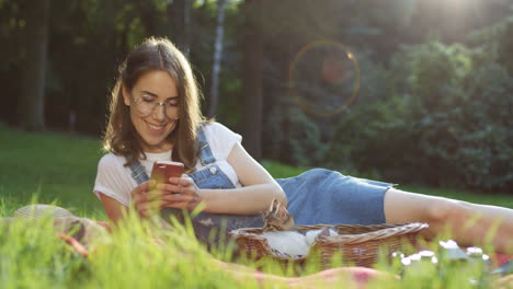 young caucasian woman in glasses lying on a blanket in the park tapping and scrolling on a smartphone