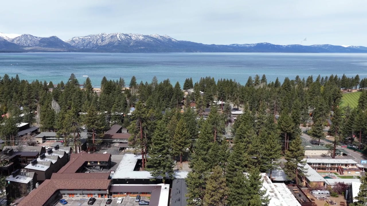 Vila turística do Lago Tahoe com chalés alpinos e densa floresta de pinheiros, lago turquesa se estendendo em direção aos picos nevados da Sierra Nevada sob céus azuis claros. Califórnia, EUA. Tomada aérea de sobrevoo