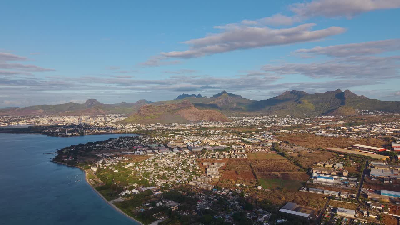 Expansive drone shot of Mauritius at sunset, revealing the island’s coastline, cityscape, and volcanic mountains bathed in golden light. Concept of tropical paradise, exploration, and island beauty