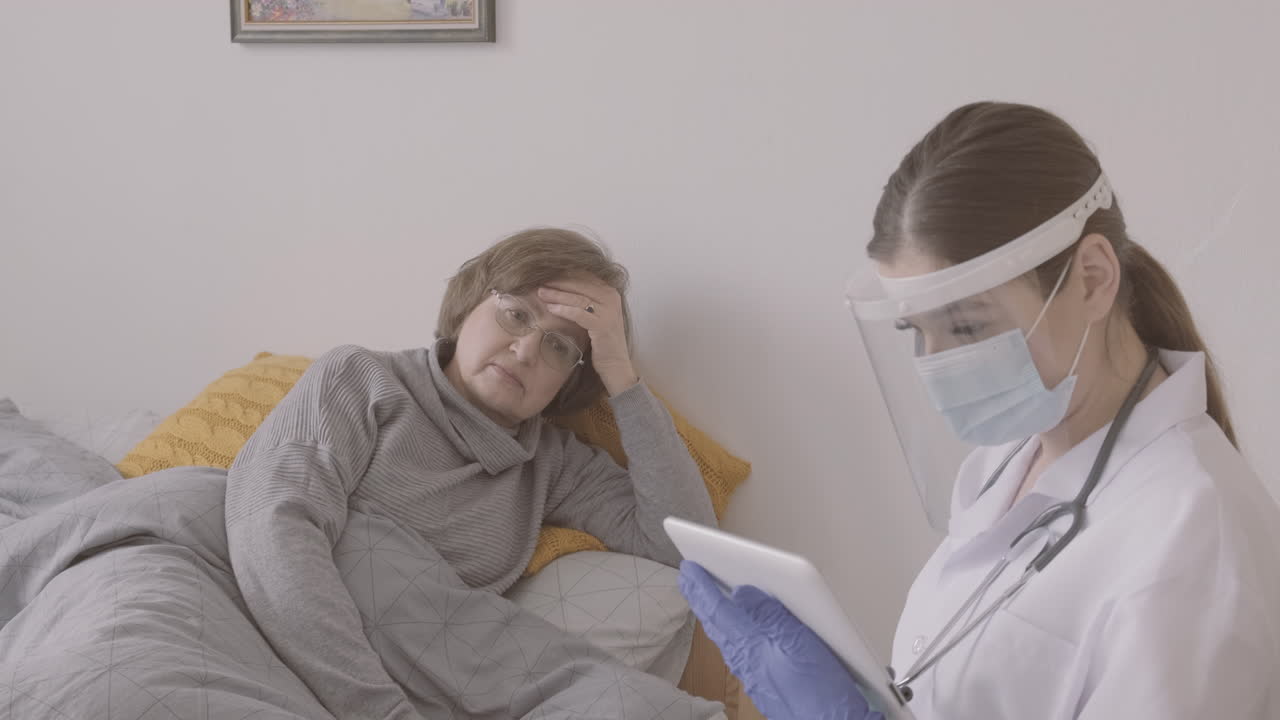 anciana sentada en la cama mientras habla con una doctora con máscara médica y pantalla protectora usando una mesa