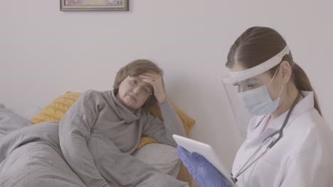 anciana sentada en la cama mientras habla con una doctora con máscara médica y pantalla protectora usando una mesa