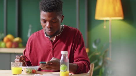 Joven-Negro-Almorzando-Y-Usando-Un-Teléfono-Inteligente-En-La-Mesa-De-La-Cocina