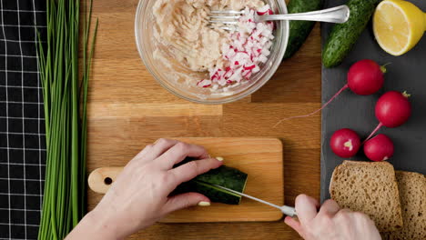 Top-down-of-amateur-cook-cutting-fresh-cucumber-into-small-chunks-to-prepare-vegetable-spread-on-wooden-kitchen-table