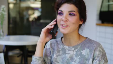 Mujer-Con-Teléfono-Inteligente-Hablando-Por-Teléfono-Mientras-Está-Sentada-En-Un-Café.-Ella-Esta-Sonriendo.-Hermosa-Mujer-Joven-Teniendo-Una-Conversación-Informal-Sobre