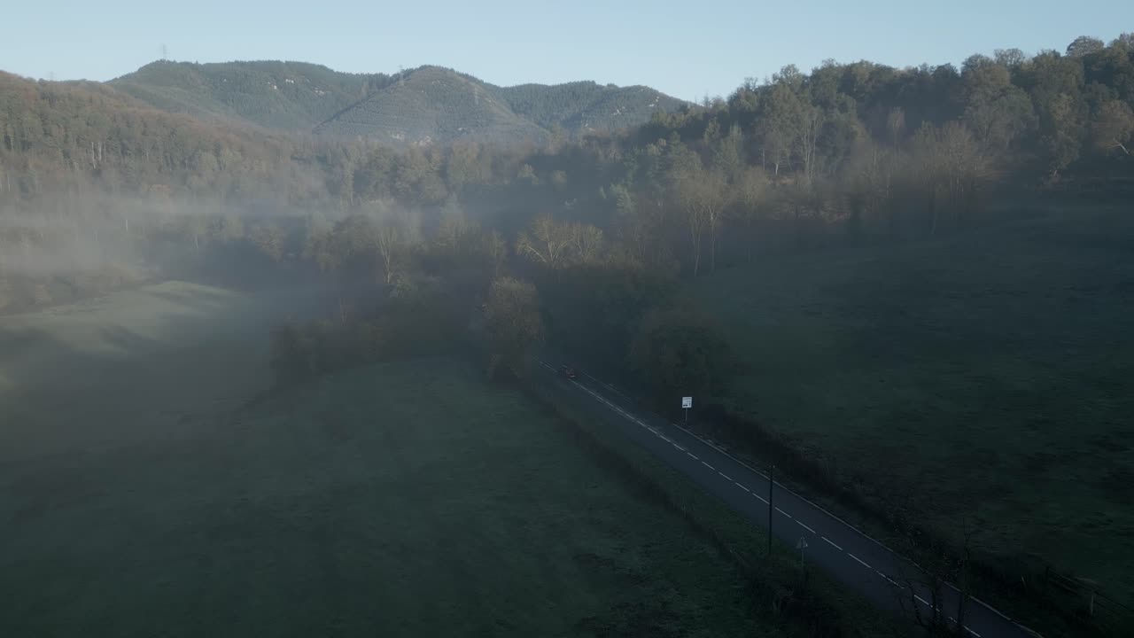 Red car driving along a two lane road that crosses a green valley surrounded by mountains and covered by fog at dawn in sant hilari sacalm, catalonia