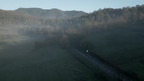 red car driving along a two lane road that crosses a green valley surrounded by mountains and covered by fog at dawn in sant hilari sacalm, catalonia