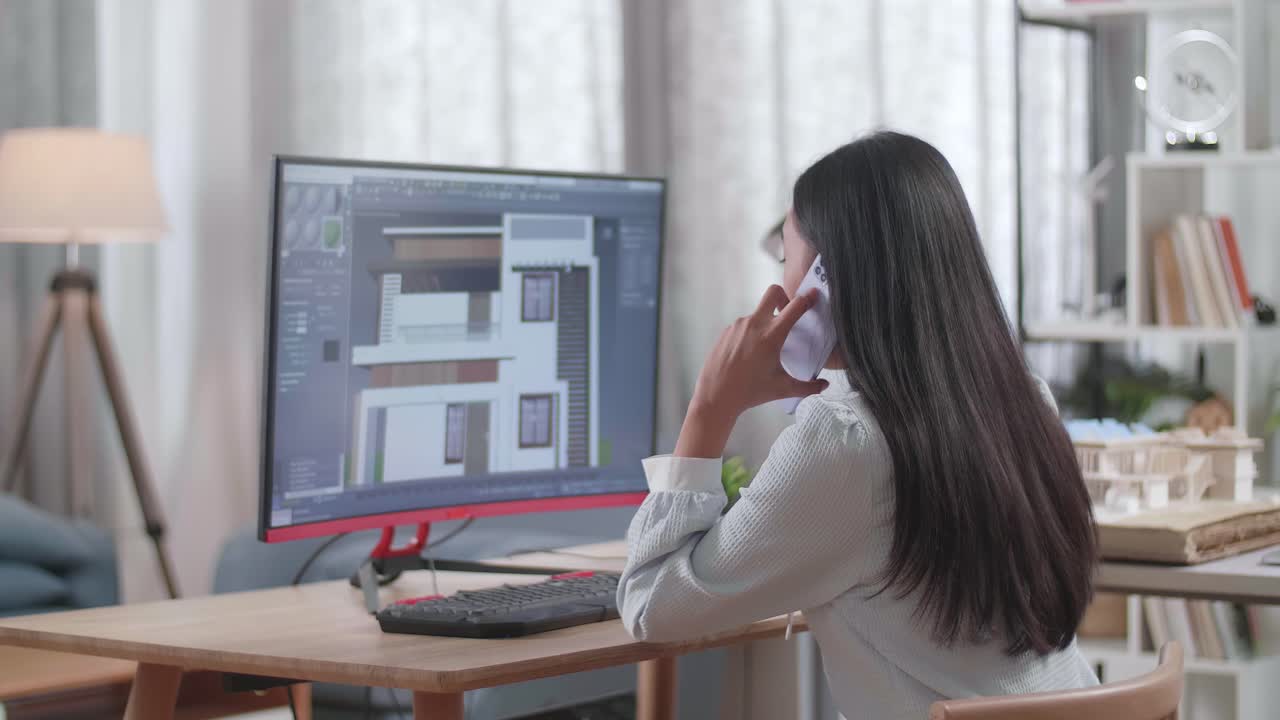 Ingegnera asiatica che parla allo smartphone mentre progetta una casa al computer a casa. Design e decorazione di case per giochi cyber
