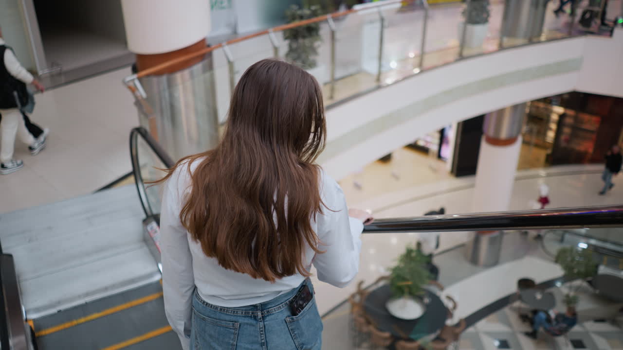 바쁜 쇼핑몰에서 레일에 손을 고 에스컬레이터를 내려가는 젊은 성인의 뒷면, 백그라운드에서 사람들이 밀링
