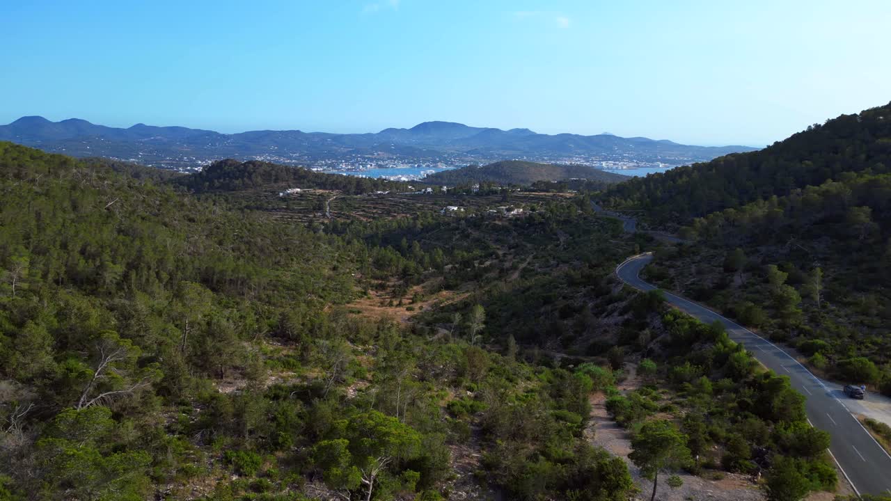 Road winding through a vibrant green forest in Ibiza, Spain, leading down towards the beautiful San Antonio Bay. Gorgeous aerial view flight fly push forward drone