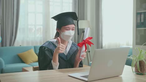 excited asian woman wearing protection face mask, thumb up and showing off a university certificate to the family during an online video call. pretty female graduate wearing a graduation gown and cap sitting on the living room