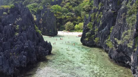 wide low aerial pull back view from hidden beach and its jungle surrounded white sand and karst rock cliffs, tourists swimming in tropical shallow turquoise water lagoon, tour c el nido, philippines