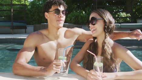 Portrait-of-happy-biracial-couple-with-drinks-at-pool-in-garden-on-sunny-day
