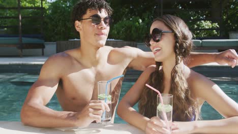retrato de una feliz pareja biracial con bebidas en la piscina en el jardín en un día soleado