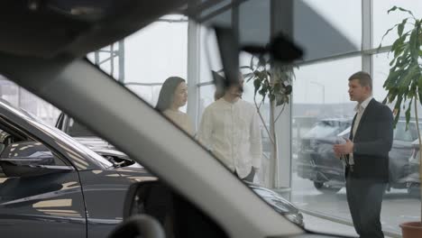 young car salesman showing to young couple new automobile at dealership salon.