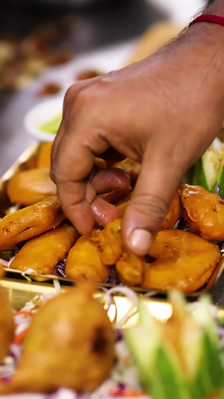 Primo piano della mano di uno chef che sistema samosa e pakora dorati su un piatto con guarnizione di verdure