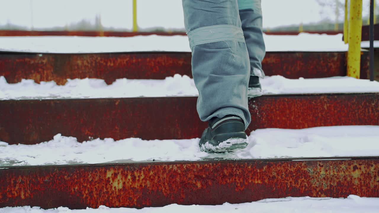 A man in a gray uniform climbs up the rusty stairs to the bridge and walks on it above the railway. Close-up on legs. Back view. Blurred background.
