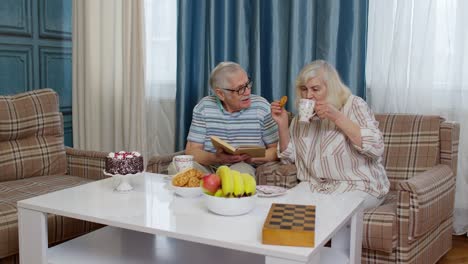 Retired-senior-couple-talking-drinking-tea,-reading-book-in-modern-living-home-room-lounge-together