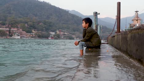 Niño-Aislado-Sentado-En-La-Orilla-De-La-Orilla-Del-Río-Santo-Ganges-Desde-Un-ángulo-Plano-Se-Toma-Un-Video-En-La-Orilla-Del-Río-Ganga-Rishikesh-Uttrakhand-India