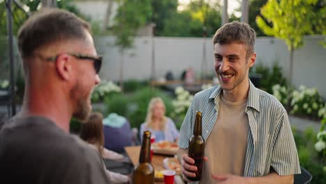 Dos-Chicos-Alegres-Charlando-Y-Divirtiéndose-Mientras-Beben-Bebidas-De-Una-Botella-De-Vidrio-Marrón-En-El-Patio-De-Una-Casa-Durante-Unas-Vacaciones-De-Fin-De-Semana.