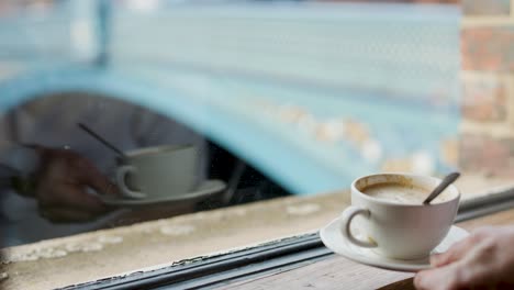 person sets coffee cup on window ledge in cafe, natural daylight, urban bridge background visible