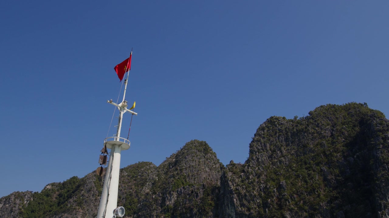 El mástil de un barco ondea la bandera vietnamita contra el cielo despejado, con karsts de piedra caliza de fondo, en la Bahía de Ha Long, provincia de Quang Ninh, Vietnam - Toma de ángulo bajo.