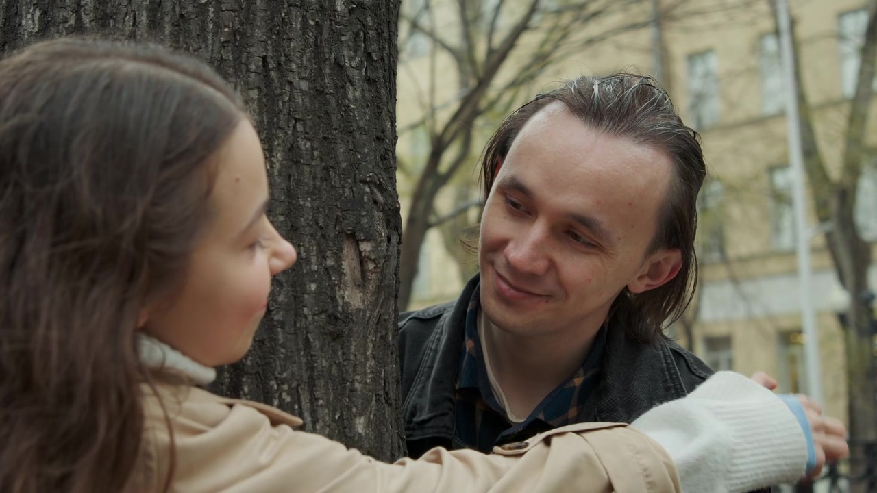 Primo piano di un uomo e una donna in piedi vicino a un albero in un parco urbano intenti a conversare. La scena presenta una luce naturale soffusa con lo skyline della città visibile sullo sfondo, inclusi edifici e alberi.