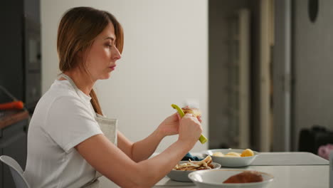 side view of woman in apron peeling potato, seated at white kitchen table, focus on hands and potato, soft blur background featuring open door, multiple bowls and peeled potatoes nearby